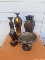 Photo showing two metal candleholders with decorative painted finishes, one metal vase with hammered finish, and metal and stone bowl centerpiece on an orange surface