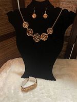 Front view of the jewelry displayed on black stands: necklace with rose designs, matching pierced earrings, and bracelet placed below.