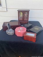 Six assorted jewelry boxes grouped on black surface outside, showing variety in size, color and style including pink glass box, shoe-shaped ring box, three black wooden boxes with gold designs, and brown wooden men’s jewelry box with glass doors and drawer.