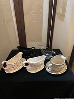 Photo showing five gravy bowls and a large clear glass serving dish on black fabric with background of brown wooden panels.