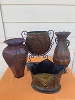 View of the four metal vases arranged on an orange cloth surface outside, showing their shapes and designs.