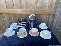 Full set of tea items including blue metal tea pot, tea tin can, plain white cups and saucers, floral print cup and saucer, and pink floral print cup and saucer on blue background with wood fence