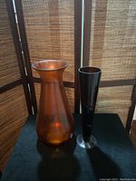 Two vintage vases on a black surface with a folding screen background, displaying an orange amber glass vase 17.5 inches tall and a tall brown swirling glass vase 15.5 inches tall.