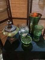 Five glass vases and dishes displayed on black cloth with wood screen background, showing green, gold, hand painted, translucent, and textured glass pieces, various shapes and sizes.