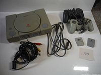 Full view of PlayStation console with two controllers, memory cards, and cables arranged around it on white surface.