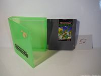 Gray NES game cartridge for Teenage Mutant Ninja Turtles with label artwork, placed next to green Nintendo cartridge storage case