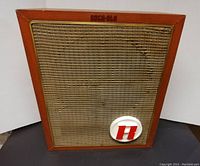 Front view showing rectangular wooden speaker with original grill cloth and Rock-Ola branding at top, as well as circular badge with red 'H'.