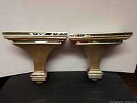 Front view showing both Art Deco style wall shelves side by side on black surface against white background.