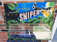 Front view of the Super Sniper arcade back glass showing the printed artwork featuring a sniper aiming through foliage and the game title.