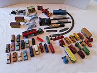 Overview of multiple die cast toy vehicles including cars, buses, planes, trains, and some accessory buildings arranged on a white surface.
