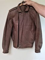 Front view of brown leather aviator style jacket with snap button collar, epaulets, and vertical pockets being held on hanger.