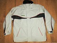 Front view of light gray waterproof winter coat with black and red details, laid flat on wooden floor