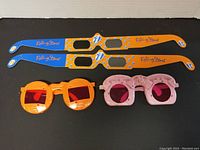 Front view of four vintage glasses showing two pairs of Rolling Stones 3D glasses with branded long rectangular arms and two pairs of Elton John style colored glasses with tinted lenses.