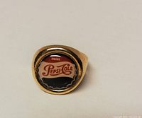 Front view of Pepsi Cola metal advertising ring featuring the bottle cap logo design in red, white, and black on a gold-tone metal setting.