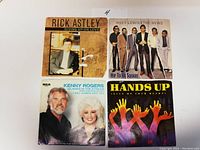 Topview photo showing fronts of four 45 RPM vinyl records with original picture sleeves: Rick Astley, Huey Lewis & The News, Kenny Rogers & Dolly Parton, Hands Up.
