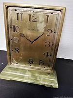 Front view of rectangular brass-faced desk clock showing the brand Tiffany & Co. and black numbers from 1 to 12 with decorative hands on a green marble base.