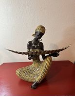 Full view of the African lady statue in a kneeling position, holding a long decorative tray on a red surface against a neutral wall.