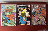 Three Action Comics and Superman-related comic books laid out on a red surface; covers show Superman in various scenes including flying and being trapped, cover prices 25 to 40 cents.