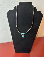 Dark beaded necklace with turquoise accent stones and a turquoise pendant displayed on black jewelry stand