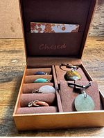 Box of five Chesed necklaces with stone pendants in blue, green, pink, reddish, and greenish stones arranged inside brown suede-lined box.