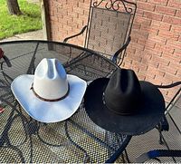 Two cowboy hats on black metal patio table; one white Arlop hat size 7 1/8 and black Cody James hat size 7 1/4