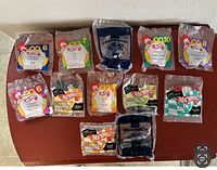 Top-down view of 12 Hot Wheels toys in original McDonald's Happy Meal packaging laid out on a table, showing various bags with different colors and numbers.