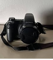 Front view of Sony Cyber-shot DSC-H5 camera with lens cap and strap visible, showing textured grip and Sony logo on lens cap.