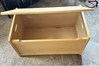 Light brown wooden toy chest with hinged lid open showing interior and side cutout handles for carrying.