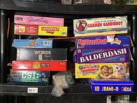 Photo showing a large variety of vintage board games stacked on a shelf including Monopoly Junior, Princess Opoly, Words, Carmen Sandiego, Rummikub, Balderdash, CSI, and others.