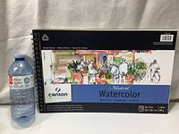 Photo of Canson Artist Series Montval watercolor paper wirebound pad next to water bottle for size reference, showing front cover with artwork and branding.