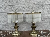 Full view of both brass table lamps with hanging glass pendants on a wooden surface against a concrete wall.