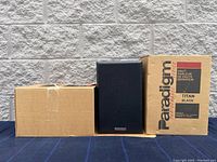 Two Paradigm Performance Series Titan Black speakers in original shipping boxes, one speaker outside box showing black mesh grill and Paradigm logo.