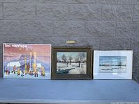 Three framed pictures arranged side-by-side: Ted Harrison exhibition poster on left, large framed original winter landscape painting in middle, smaller framed winter landscape painting on right.