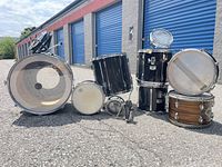 Full kit overview displaying bass drum, rack toms, floor tom, snare drums and hardware components on pavement outside storage units
