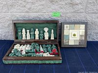 Photo of heavy chess set inside an open wood frame case next to the tile wall decor, all placed on a dark fabric with a concrete wall behind.