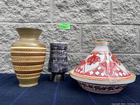 Three ceramic pieces on a blue cloth backdrop against a stone wall: large beige vase with patterned bands, small blue-black textured vase, colorful ceramic tagine dish with lid