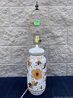 Full view of floral decorated ceramic lamp showing tall white speckled ceramic base with orange, yellow, olive, and brown flowers and black vine detailing, brass socket, and lamp harp.