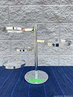 Chrome swivel plant holder shown from multiple angles against a stone background on a dark cloth. Shows four circular rings for plants mounted on swivel arms around a central chrome pole with a round base.