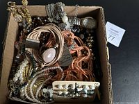 Top-down image of a cardboard box filled with assorted vintage jewelry including necklaces, bracelets, earrings, and a wristwatch.
