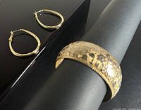 A close-up photo showing a gold tone cuff bracelet with intricate decoration and a pair of gold tone oval hoop earrings placed on a black surface.