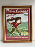 Framed vintage advertisement poster showing a lumberjack carrying a log with a scenic forest background and promotional text for British Columbia and Strathcona Park.