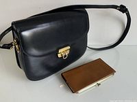 Photo shows a black leather shoulder bag with a flap and gold-tone clasp, alongside a brown leather wallet with a metallic clasp.