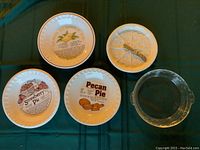 Photo showing all six plates on table: one clear Pyrex pie plate, three recipe printed ceramic pie plates (lemon meringue, strawberry, pecan), one white pressed shortbread plate with small metal pie server.