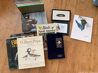 Photo showing a collection of bird and mammal books, including 'Birds of North America', 'William Pope', and 'The Encyclopedia of Mammals', with framed bird art and a painting on a feather beside them.