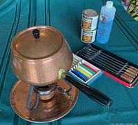 Copper fondue pot with black handle and lid knob, two sets of fondue forks, two small fuel cans and blue liquid bottle around it on green surface.