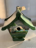 Handpainted wooden birdhouse front with circular entrance hole and scenic artwork under green roof