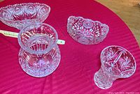 Four clear heavy crystal pieces on a red tablecloth including two oval bowls and two vases with intricate cut patterns.
