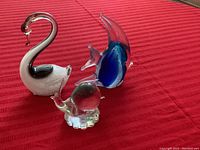 Three small glass figurines on a red textile surface: a swan, an angel fish, and a snail, showing minor chips.