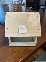 White wooden bird feeder with sloped roof and hanging wire shown on a wood table.