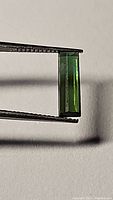 Close-up photo showing rectangular baguette cut green tourmaline gemstone held with tweezers, highlighting color, cut and clarity.